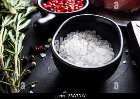 Sel de mer dans un bol noir et herbes de romarin gros plan sur la table en pierre noire avec épices et viande crue près de la vue latérale sélective. Banque D'Images