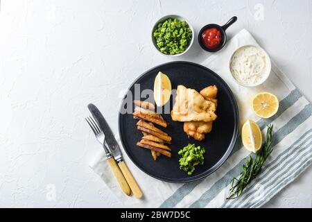 Fish and chips traditionnel britannique avec détail petits pois et une tranche de citron. Sur plaque ronde noire sur lintn blanc et surface de pierre Banque D'Images