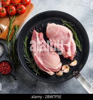 Steak de porc sur un os dans une poêle à frire noire sur fond de pierre texturée grise avec un tissu noir romarin ail grains de poivre et tomates sur la hache Banque D'Images