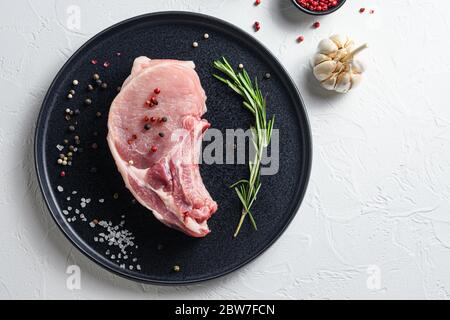 Steak de porc frais dans une assiette ronde noire sur fond de pierre blanche avec des ingrédients de poivre de romarin ail pour le gril espace de vue de dessus pour le texte. Banque D'Images