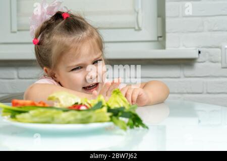 une petite fille repousse émotionnellement une assiette de légumes offerts. les enfants n'aiment pas les légumes Banque D'Images