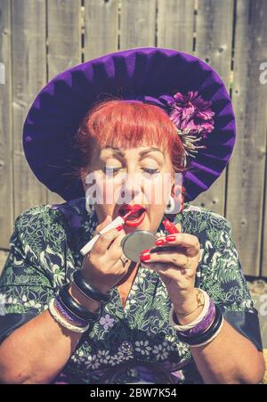 Gros plan de la femme isolée en BID, chapeau violet mettant sur rouge à lèvres brillant, à l'extérieur sous le soleil à l'événement de reconstitution 1940s WW2, Royaume-Uni. Banque D'Images