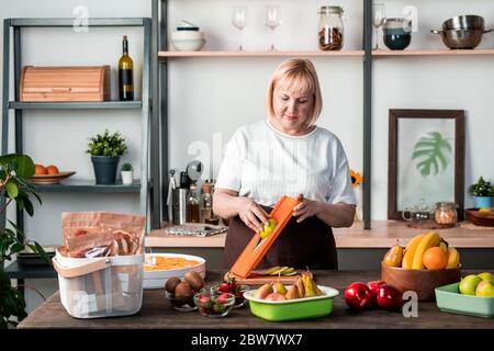 Blonde femme en tenue décontractée debout près de table de cuisine avec variété de fruits frais et à l'aide d'un couteau tout en préparant des poires faites maison sèches Banque D'Images