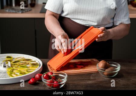 Mains de femme au foyer contemporain avec couteau hachant des fraises fraîches sur une planche en bois pour les mettre sur plateau de sèche-fruits Banque D'Images