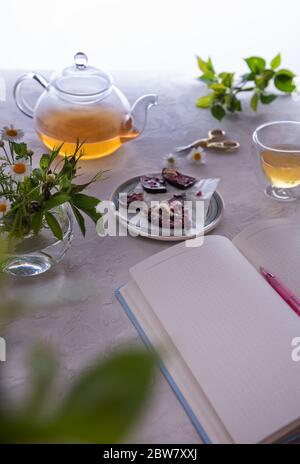 Journal ouvert à une page vide avec un stylo rose. Il y a aussi une théière en verre non concentrée, une tasse thermo avec thé, des chocolats et un bouquet de fleurs dans un vase. Banque D'Images