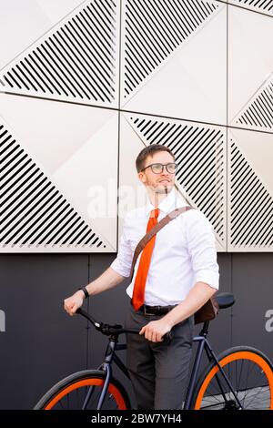 Jeune directeur de bureau réussi en chemise, cravate et pantalon regardant de côté et tenant les poignées de vélo tout en se tenant contre l'extérieur du bâtiment Banque D'Images