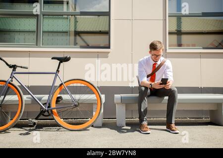 Messagerie élégante pour jeunes hommes dans un téléphone intelligent, assis sur un banc contre le mur et les fenêtres du centre d'affaires contemporain Banque D'Images