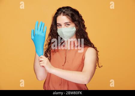 Jeune femme sérieuse jolie en chemise et masque de protection mettant des gants bleus pour éviter l'infection à covid19 en période de quarantaine Banque D'Images