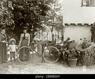 Un homme pose sur son vélo (en équilibre avec l'aide d'un pot de fleur retournée) tandis que d'autres membres de sa famille regardent, Gloucestershire, Angleterre, Royaume-Uni c. 1900. L'homme porte un costume et un chapeau de paille, tout comme une dame et un petit garçon. Les personnes représentées sont des travailleurs agricoles. Ils étaient également des jardiniers enthousiastes avec leurs plantes en pot en démonstration. Notez l'utilisation de troncs d'arbres creusés pour fabriquer des jardinières. Banque D'Images