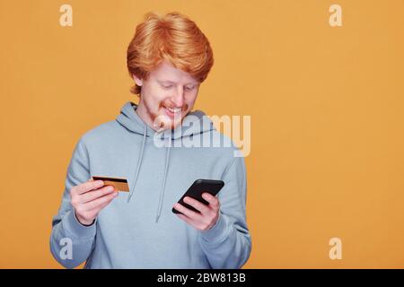 Joyeux jeune homme décontracté avec smartphone défilant à travers les marchandises dans la boutique en ligne et allant payer par carte de crédit tout en faisant la commande Banque D'Images