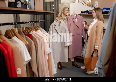 Bonne jeune femme blonde montrant deux manteaux élégants de la collection saisonnière à sa mère tout en choisissant de nouveaux vêtements dans le centre commercial Banque D'Images