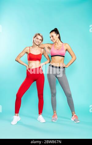 Prêt pour l'entraînement. Deux jeunes filles sportives attrayantes et séduisantes en vêtements de sport, en regardant l'appareil photo et en souriant, debout contre le bleu Banque D'Images