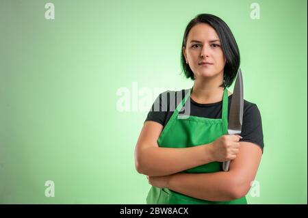 Une femme cuisinait avec un tablier vert et un t-shirt noir, en regardant avec confiance le couteau dans sa main isolé sur fond vert Banque D'Images
