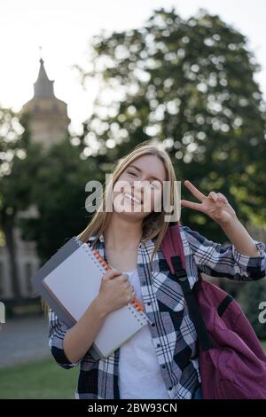 Fille gaie montrant le signe de victoire. Concept de retour à l'école. Portrait d'un étudiant souriant avec sac à dos et livres à pied à l'université Banque D'Images