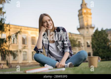 Fille de collège assise sur l'herbe dans le parc. Concept d'éducation. Portrait d'une jeune femme attrayante regardant l'appareil photo et riant Banque D'Images