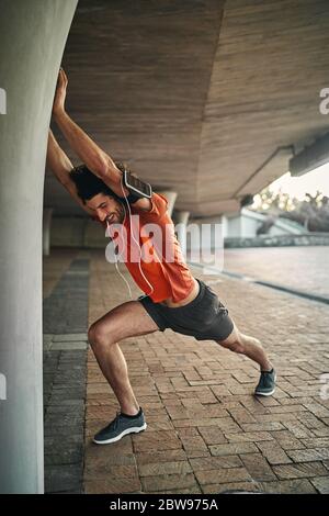 Vue latérale d'un jeune homme de forme physique joyeux avec des appareils électroniques qui lui permettent de se faire passer la main sur la colonne sous le pont Banque D'Images