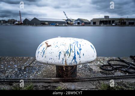 Un bollard blanc avec des taches de peinture sur un quai avec des maisons de guerre en arrière-plan. Ciel sombre. Prise de vue en exposition longue. Banque D'Images