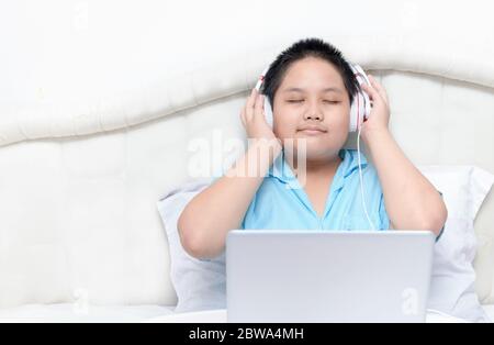Mignon garçon asiatique heureux portant des écouteurs pour écouter de la musique depuis un ordinateur portable sur le lit dans la chambre. Concept d'éducation et de technologie Banque D'Images
