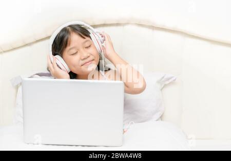 Adorables filles asiatiques portant des écouteurs pour écouter de la musique depuis un ordinateur portable sur le lit de la chambre. Concept éducatif et technologique Banque D'Images