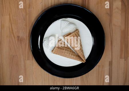 dessert blanc rond gâteau doux et savoureux à la crème glacée avec décoration deux cônes de glace dans une tasse à gaufres sur une assiette noire sur fond de bois. en haut Banque D'Images