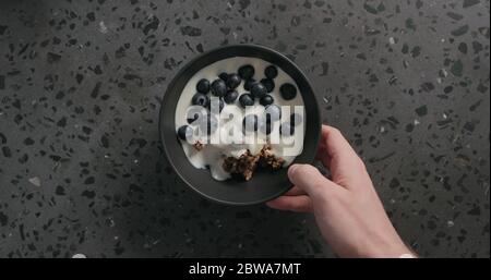 vue de dessus les mains de l'homme mettre le granola de chocolat, les flocons d'amande et le myrtille dans un bol noir sur la surface de terrazzo Banque D'Images