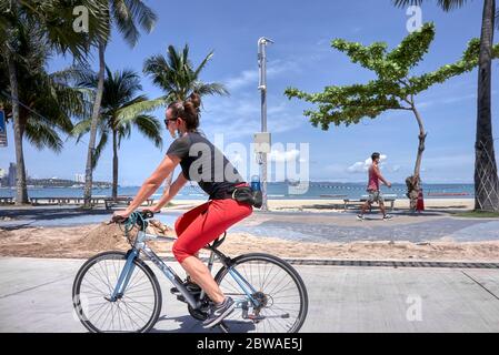 Exercice Covid 19. Femme prenant l'exercice de vélo pendant le verrouillage du coronavirus avec l'homme marchant dans l'arrière-plan. Thaïlande, Asie Banque D'Images