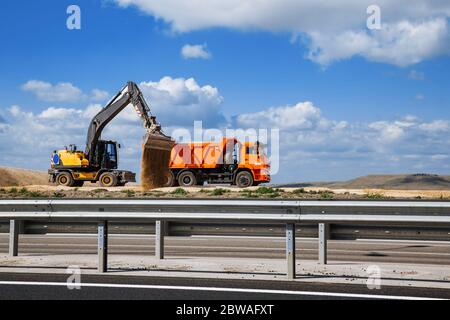 Une pelle hydraulique jaune verse le sable d'un gros camion sur la route avec un godet. Construction de routes. Banque D'Images