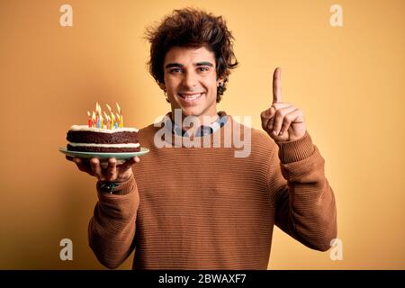 Jeune homme beau tenant un gâteau d'anniversaire debout sur fond jaune isolé surpris par une idée ou une question pointant du doigt avec le visage heureux, Banque D'Images