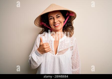 Femme brunette d'âge moyen portant un chapeau conique traditionnel asiatique sur fond blanc faisant des pouces heureux vers le haut geste avec la main. Approbation de l'expression lo Banque D'Images