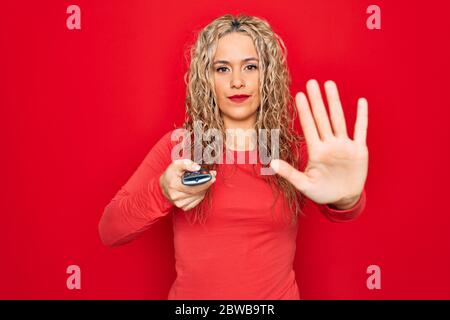 Jeune belle femme blonde utilisant la télécommande de télévision sur fond rouge isolé avec main ouverte faisant signe d'arrêt avec sérieux et confiant ex Banque D'Images