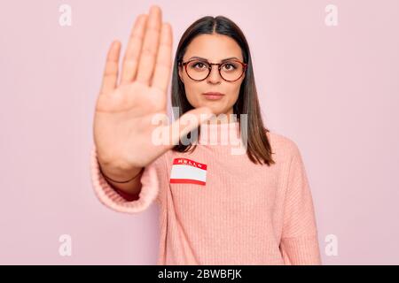 Jeune belle femme brunette portant l'autocollant avec bonjour mon nom est message avec main ouverte faisant stop signe avec l'expression sérieuse et confiante, def Banque D'Images