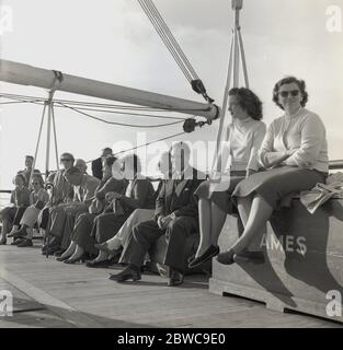 Dans les années 1950, les passagers adultes historiques naviguant sur un navire à vapeur Union-Castle se sont dirigés vers le Cap en Afrique du Sud et ont les vêtements formels de l'époque, assis à l'extérieur sur le bord du pont, attendant peut-être de débarquer du bateau. Deux dames sont assis sur la boîte en bois tenant les jeux de plate-forme. Union-Castle était une ligne de navigation britannique qui exploitait des navires mixtes de passagers et de courrier entre l'Europe et l'Afrique de 1900 à 1977. Banque D'Images