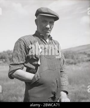 Années 1950, photographie historique de cette époque d'un fermier de tourbe irlandais qui déversait des salopettes de coton déchirées et une casquette de tissu debout par des blocs de tourbe qu'il vient de creuser, Irlande. Dans les pays ruraux comme l'Irlande, la tourbe était et reste une ressource précieuse utilisée comme combustible pour la cuisine et le chauffage domestique pour ceux qui vivent hors de la terre comme les agriculteurs et les petits exploitants. Banque D'Images