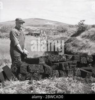 Années 1950, photographie historique de cette époque d'un fermier de tourbe irlandais qui déversait des salopettes de coton déchirées et une casquette de tissu debout par des blocs de tourbe qu'il vient de creuser, Irlande. Dans les pays ruraux comme l'Irlande, la tourbe était et reste une ressource précieuse utilisée comme combustible pour la cuisine et le chauffage domestique pour ceux qui vivent hors de la terre comme les agriculteurs et les petits exploitants. Banque D'Images