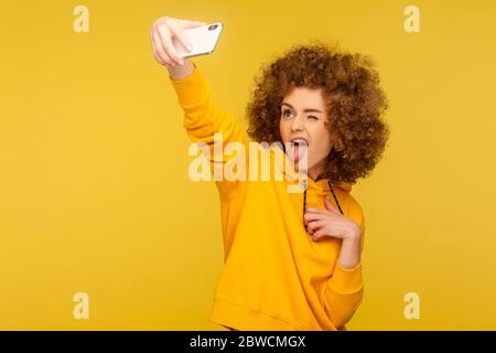 Portrait de la femme à cheveux bouclés sans souci dans un sweat à capuche de style urbain prenant le selfie et grimaquant avec la langue dehors, s'amuser tout en faisant la photo, vlog br Banque D'Images
