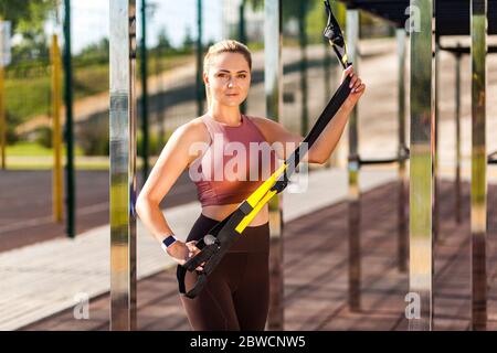 Belle femme sportive en collant sport entraînement en extérieur jour d'été, debout avec des sangles de fitness trx et regardant l'appareil photo avec motivation confid Banque D'Images