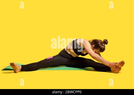 Femme mince de gymnastique avec cheveux de pain dans les vêtements de sport serrés assis sur le tapis avec les jambes étalées, se pencher pour toucher les orteils, faire des exercices d'étirement de muscle, flexibil Banque D'Images