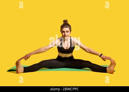 Femme de gymnastique avec cheveux de pain dans les vêtements de sport serrés assis sur le tapis avec les jambes étalées, touchant les orteils étirant les muscles, faisant des exercices de flexibilité sportive. Banque D'Images