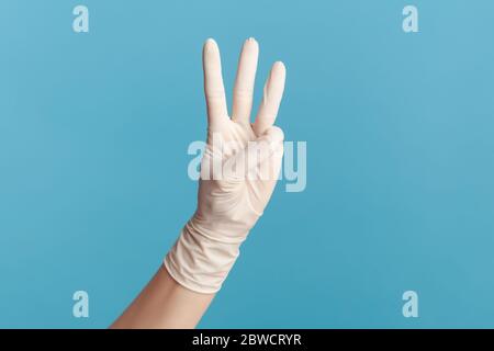 Vue latérale du profil gros plan de la main humaine avec des gants chirurgicaux blancs portant le numéro 3 trois avec les mains. Vue en salle, prise en studio, isolée sur fond bleu. Banque D'Images