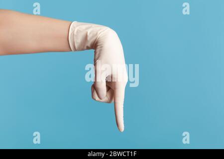 Vue latérale du profil gros plan de la main humaine avec des gants chirurgicaux blancs montrant ou pointant vers le bas avec le doigt. Vue en salle, en studio, isolée sur le dos bleu Banque D'Images