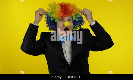 Clown caucasien senior dans la perruque colorée et noeud bleu faisant des visages stupides, idiot autour. Femme âgée s'amusant, souriant, regardant la caméra. Copier l'espace. Halloween. Fond jaune Banque D'Images