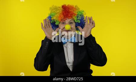 Clown caucasien senior dans la perruque colorée et noeud bleu faisant des visages stupides, idiot autour. Femme âgée s'amusant, souriant, regardant la caméra. Copier l'espace. Halloween. Fond jaune Banque D'Images