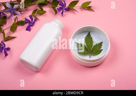 bouteilles blanches avec cosmétiques pour le corps avec feuille de marijuana sur fond rose avec fleurs sauvages violettes. soins de la peau, beauté, cosmétiques avec cannabis Banque D'Images