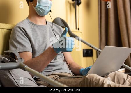 Concentration sélective de l'homme dans le masque médical et le casque montrant le pouce vers le haut pendant l'appel vidéo sur l'ordinateur portable près des béquilles sur le lit Banque D'Images