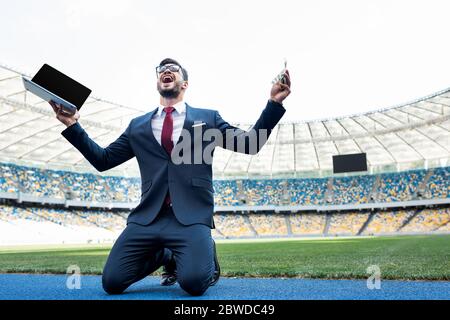 un jeune homme d'affaires heureux en costume tenant un ordinateur portable avec écran vierge et de l'argent tout en se tenant à genoux au stade Banque D'Images