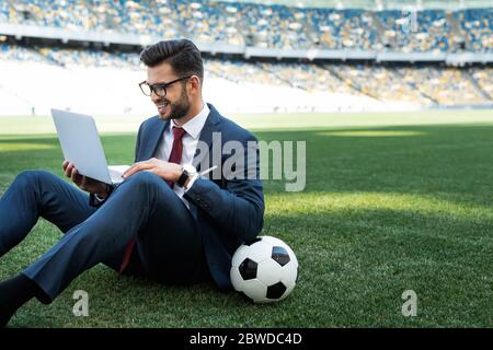 jeune homme d'affaires souriant en costume avec ordinateur portable et ballon de football assis sur le terrain de football au stade, concept de paris sportifs Banque D'Images