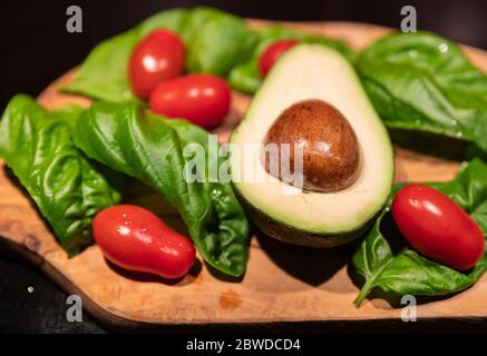 Une planche à découper en bois sur fond noir avec un avocat au centre. Tout autour, feuilles de basilic vert intense et tomates cerises rouges brillantes. Banque D'Images