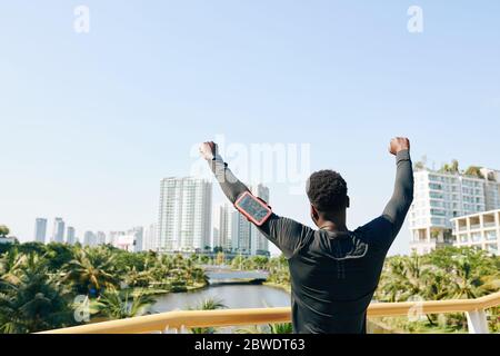 Un sportif enthousiaste qui lève les bras et regarde la ville depuis le point culminant de sa course matinale, vue de l'arrière Banque D'Images