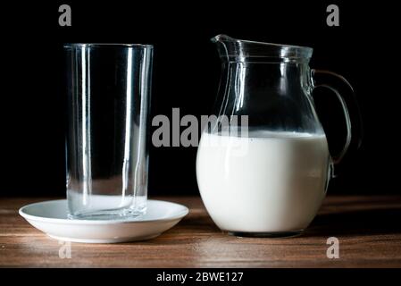 Une tasse en verre vide et un pot de lait de vache sur une table en bois à fond noir Banque D'Images