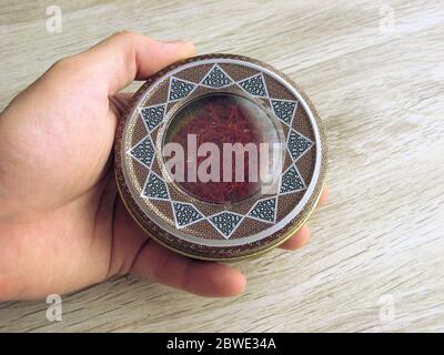 Mashhad, IRAN, 10 03 2020: Boîte contenant du safran rouge dans la main mâle sur fond de bois. Souvenir iranien. L'épice le plus cher. Banque D'Images